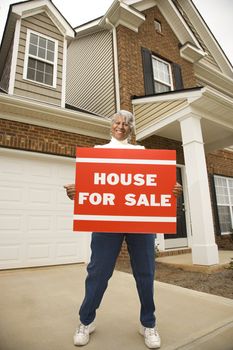 Portrait of middle-aged African-American female outside house with for sale sign.