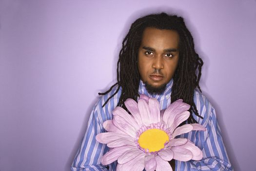 Serious African-American mid-adult man holding big fake flower on purple background.