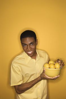 Smiling young African-American man holding bowl of lemons on yellow background.