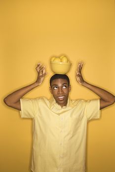Young African-American man balancing bowl of lemons on his head on yellow background.