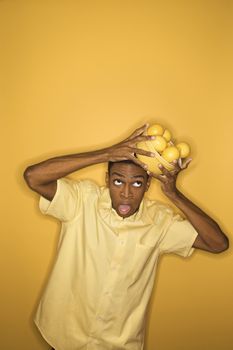 Young African-American man dropping a bowl of lemons balancing on his head, on yellow background.