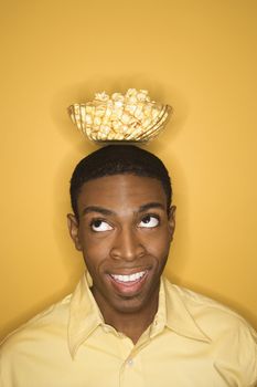 Young African-American man balancing bowl of popcorn on his head on yellow background.