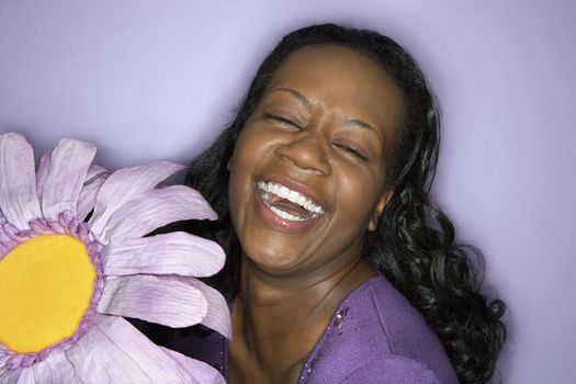 Portrait of African-American mid-adult woman smiling big with eyes closed holding big purple fake flower on purple background.