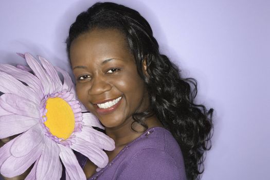 Portrait of smiling African-American mid-adult woman holding big purple fake flower on purple background.