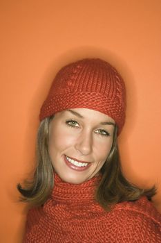 Head and shoulder portrait of smiling young adult Caucasian woman on orange background wearing winter hat and scarf.