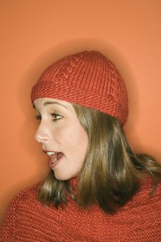 Side view of young adult Caucasian woman on orange background wearing winter hat and scarf.