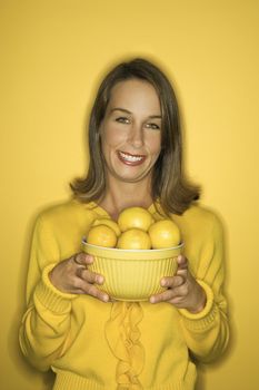 Portrait of smiling young adult Caucasian woman on yellow background holding bowl of lemons.