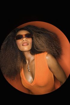 Vignette of young African-American adult woman wearing sunglasses with big hair and low cut dress on orange background seductively looking at the viewer.