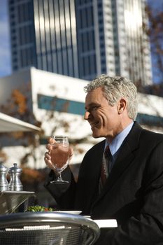 Prime adult Caucasian man in suit sitting outside drinking water at patio table in urban setting.