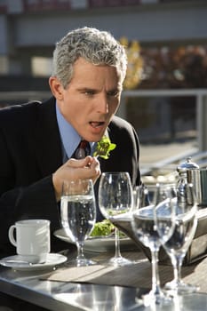 Prime adult Caucasian man in suit sitting at outside patio table eating salad.
