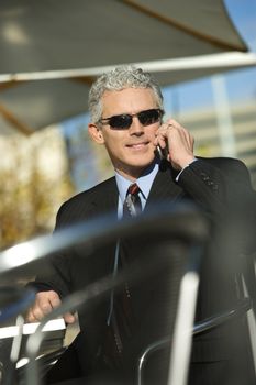 Prime adult Caucasian man in suit sitting at ouside patio table wearing sunglasses and talking on cellphone smiling.