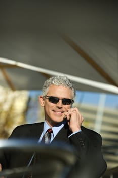 Close up of prime adult Caucasian man in suit sitting at patio table outside talking on cellphone and smiling.