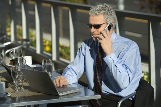 Prime adult Caucasian man in suit sitting at patio table outside looking at laptop and talking on cellphone.