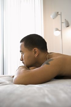 Profile of shirtless Asian young adult man lying on bed resting head on arms looking out window.