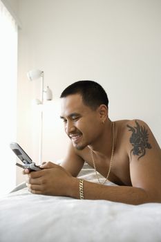 Smiling shirtless Asian young man lying on bed dialing or text messaging on cellphone.