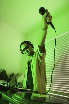 Smiling Asian young adult male DJ wearing sunglasses and holding microphone up in front of mixing equipment and turntable.