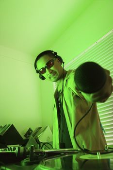 Asian young adult male DJ standing behind mixing equipment and turntable holding microphone out to viewer.