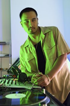 Asian young adult male DJ leaning on turntable looking at viewer.