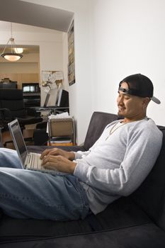 Asian young adult man sitting on sofa wearing cap backwards and typing on laptop.