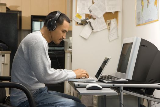 Side view of Asian young adult man sitting at desk typing on laptop wearing headphones.