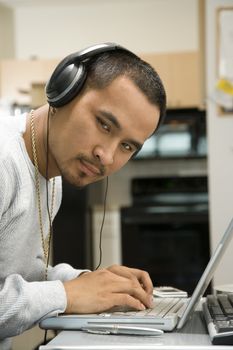 Close-up of Asian young adult man wearing headphones working on laptop and looking at viewer.