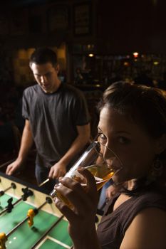 Young woman drinking beer and looking at viewer while man plays foosball in pub.
