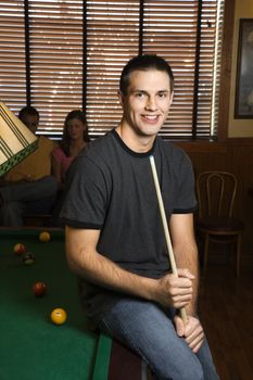 Portrait of young man leaning on billiards table holding pool stick.