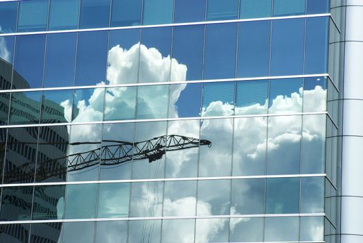 Image of a metallic crane reflected in a building's glass panels. White clouds and blue sky in the background.