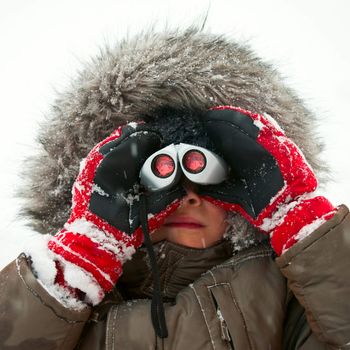 Young boy wearing winter jacket with furry hood and red gloves looking through binocular