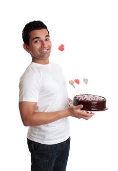 Cheeky flirtatious man carries a chocolate cake decorated with love hearts.    He has one heart clenched between his teeth.  Valentines, birthday or special occasion.  Side view against a white backdrop.