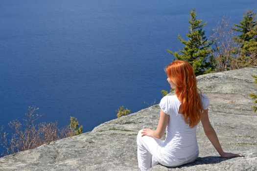Red-haired girl sitting on a rock, enjoying the view over lake.