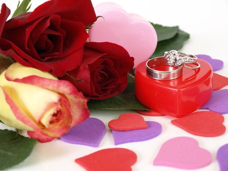 Wedding rings sit atop a red plastic box. Three roses lay next to it, various heart shapes scattered around.