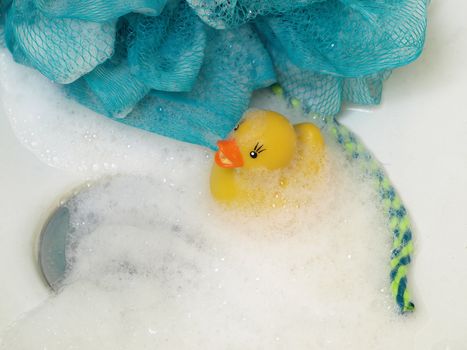 A yellow rubber duck sits near a tub drain in bubbles. A soapy scrub cloth in the background.