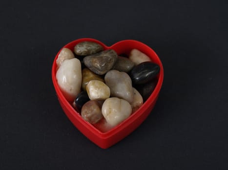 A red plastic heart shaped container holds a variety of polished decorative stones. Over black.