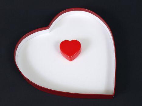 A red plastic heart sits alone inside a white heart shaped box over a black background.