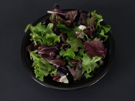 A black plate of salad with green and purple leaves over a black background.