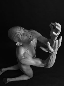 A bald and nude male on his knees, pleading, against a black background. In black and white.