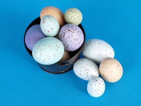Speckled pastel colored eggs fill a metal pail, a few eggs laying to the side, over a blue background.
