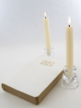 A white Holy Bible between two white lit candlesticks on a white background.