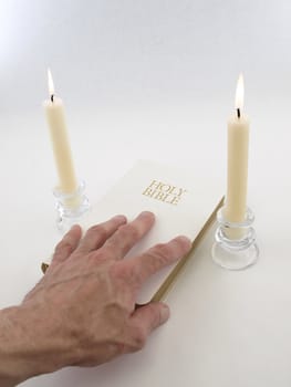 A man's hand touches the cover of a white Holy Bible. Two taper candles burn in the background. 