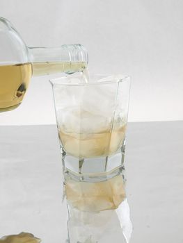 A bottle of scotch being tipped into a glass full of ice over a reflective surface.