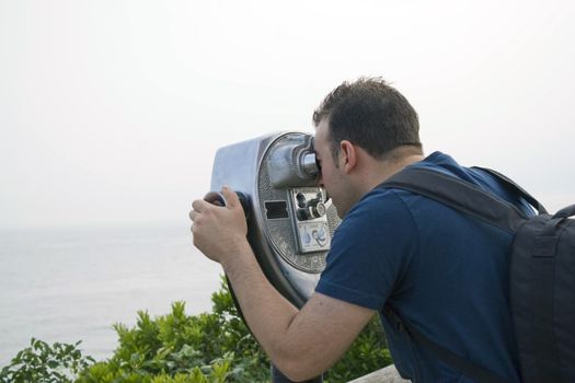 A young man looks through some coin-operated binoculars at the sea shore.