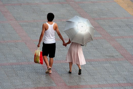 Young adult man and woman walking together in the street. The umbrella of the woman contrast with the clothes of the man but it is because the umbrella protect against the sun. Typical street scene in China. I take this picture in Xi'an.