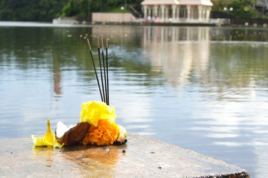 Offerings of sandal sticks, fruits and flowers during a prayer on the shore of a lake. Temple in the background.