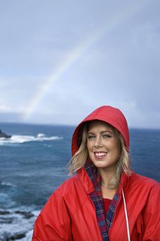 Caucasian mid-adult woman in red raincoat in front of ocean with rainbow in background looking at viewer and smiling in Maui, Hawaii.