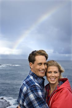 Caucasian mid-adult couple standing by ocean with rainbow in background in Maui, Hawaii.