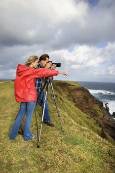 Caucasian mid-adult couple pointing and looking through camera from cliff overlooking the ocean in Maui, Hawaii.