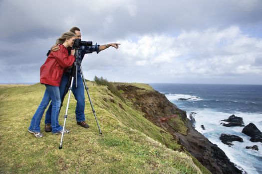 Caucasian mid-adult couple pointing and looking through camera from cliff overlooking the ocean in Maui, Hawaii.