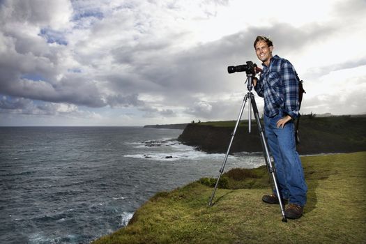 Caucasian mid-adult male standing with camera on tripod looking at viewer on cliff overlooking the ocean in Maui, Hawaii.