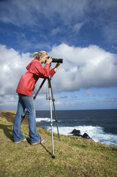 Caucasian mid-adult woman looking through camera on tripod on cliff overlooking ocean in Maui, Hawaii.